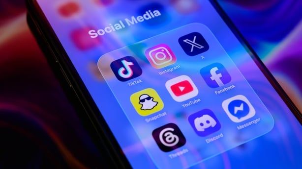 A folder of social media applications, including TikTok, Instagram, X (formerly Twitter), Snapchat, YouTube, Facebook, Threads, Discord, and Messenger, is displayed on a smartphone screen placed on a reflective surface onto which a colorful abstract illustration is projected, in Creteil, France, on