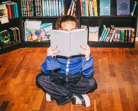 A person sits cross-legged on a wooden floor, holding an open book covering their face, in front of bookshelves