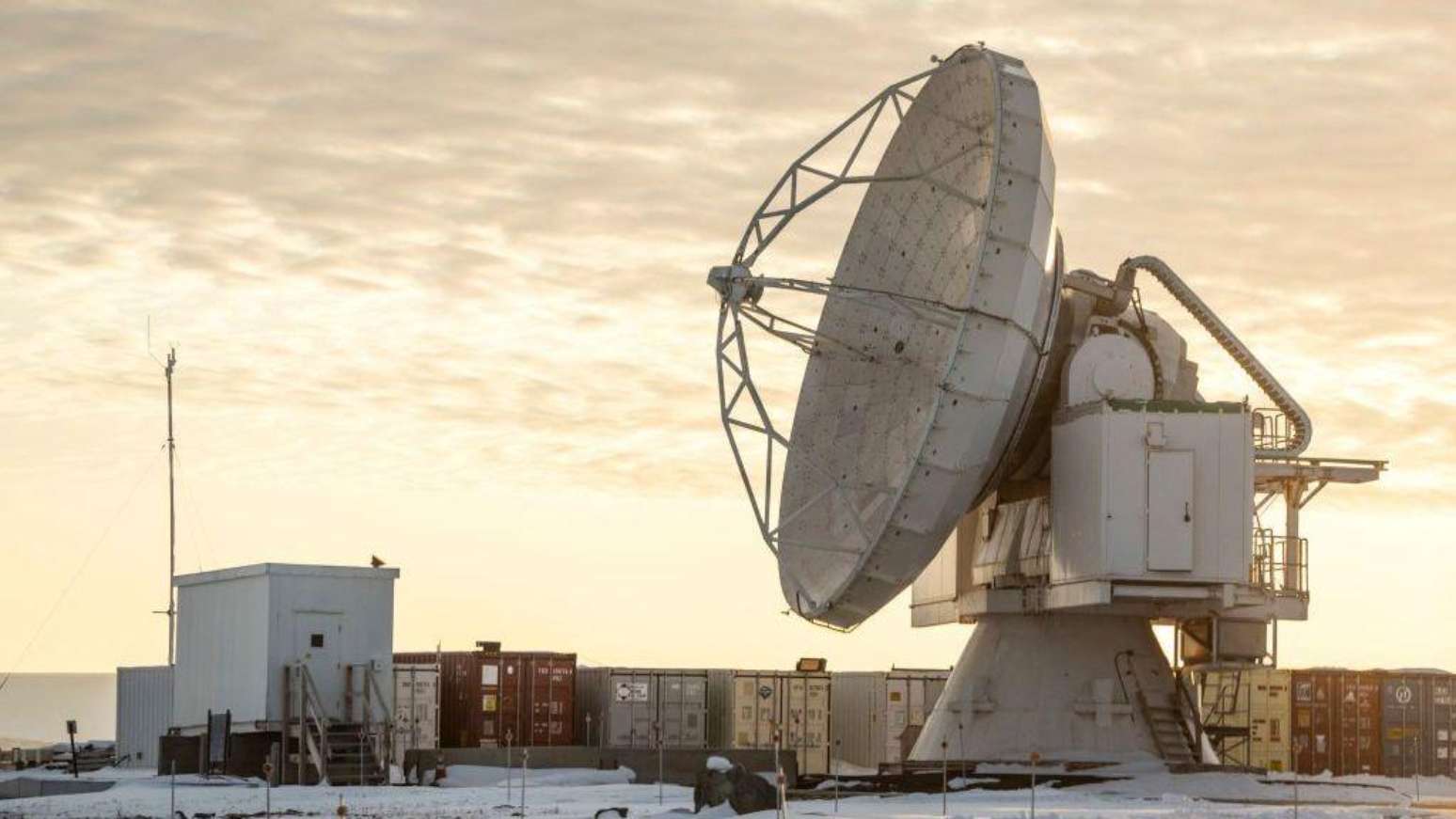A Base Espacial Pituffik (antiga Base Aérea de Thule) é mostrada no norte da Groenlândia, em 4 de outubro de 2023. A imagem mostra uma grande antena parabólica em frente a contêineres, com um céu alaranjado e nublado ao fundo.
