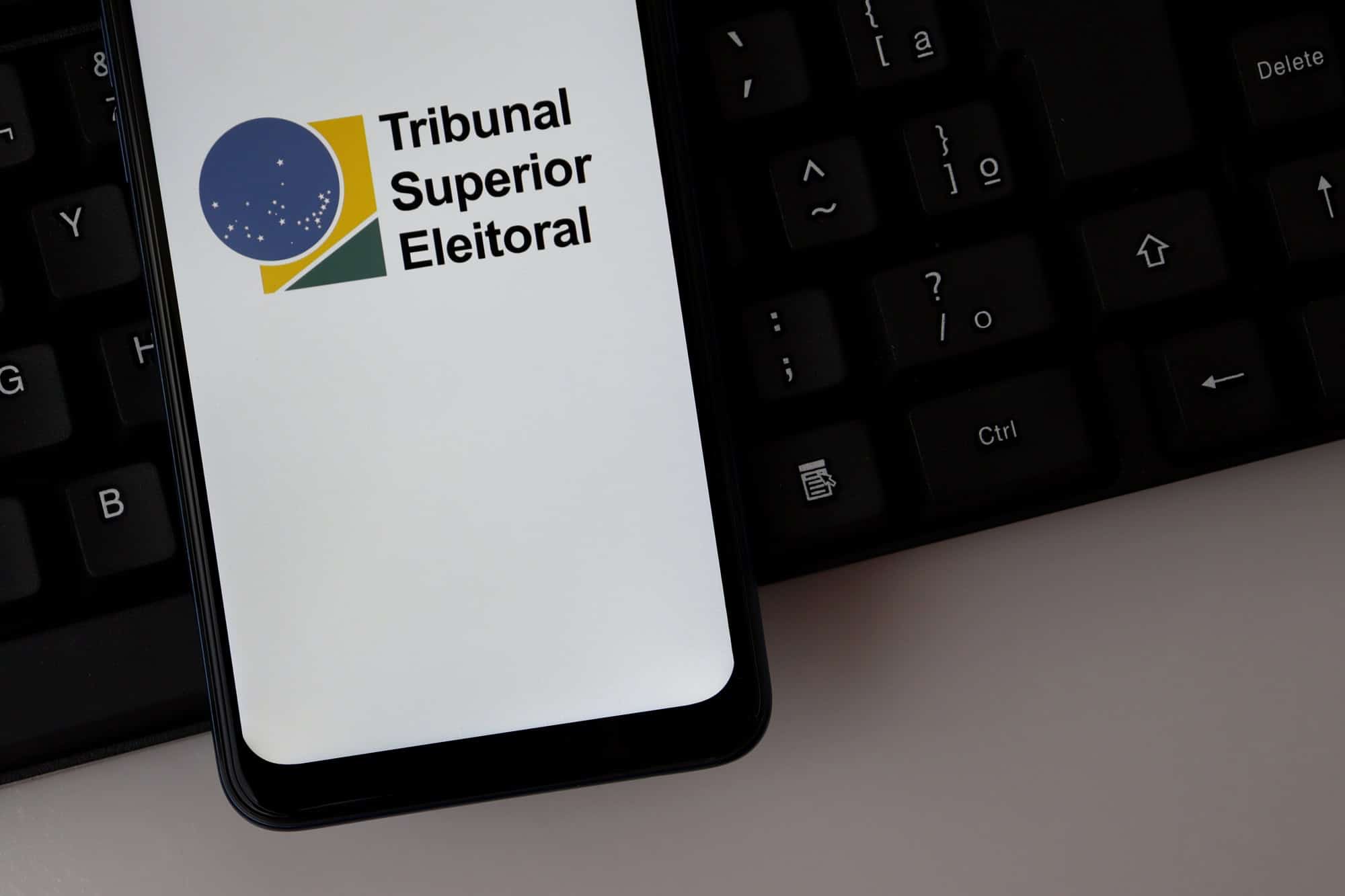 Logotipo do Tribunal Superior Eleitoral (TSE) exibido na tela de um smartphone sobre um teclado de computador.