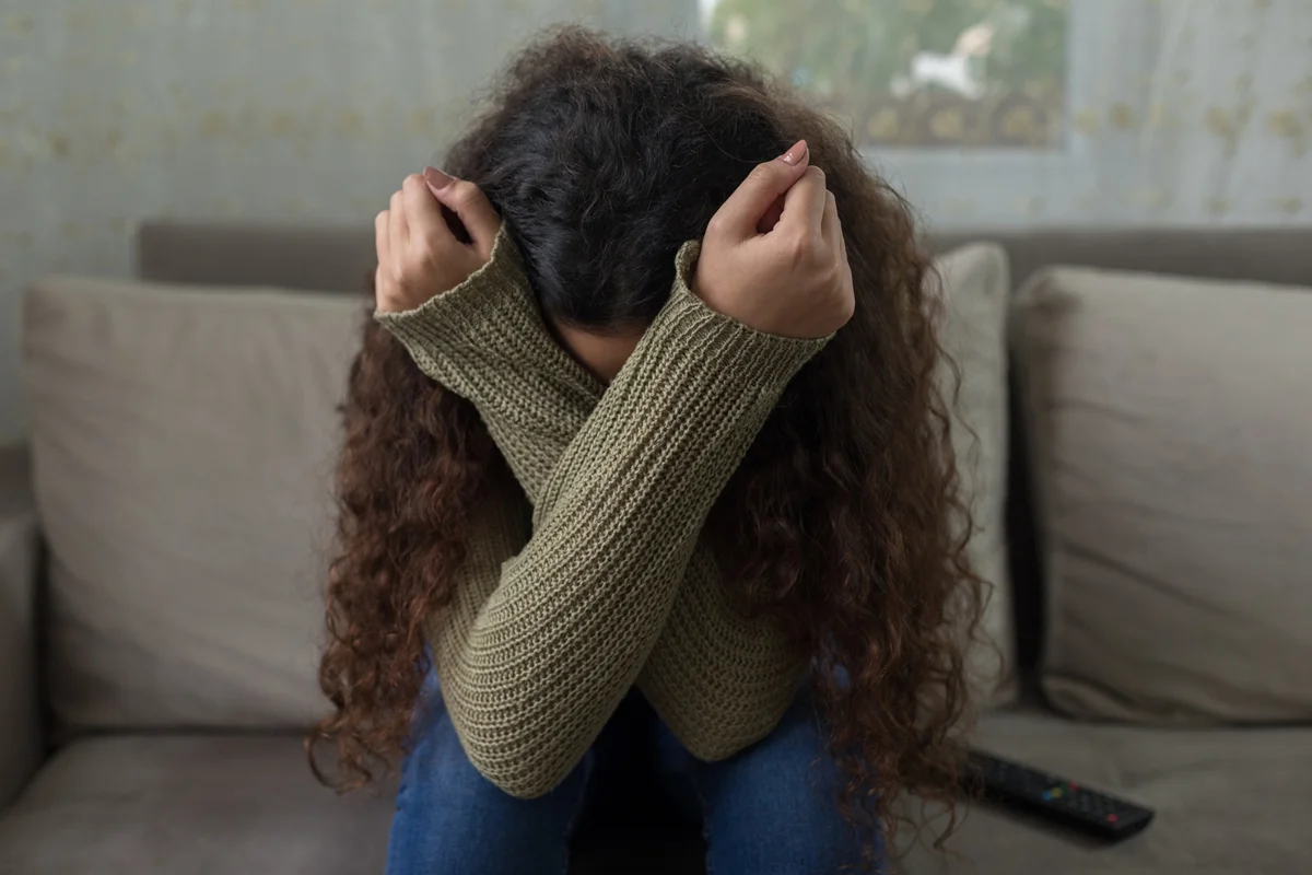 Imagem de uma menina triste sentada no sofá cobrindo o rosto. Metrópoles